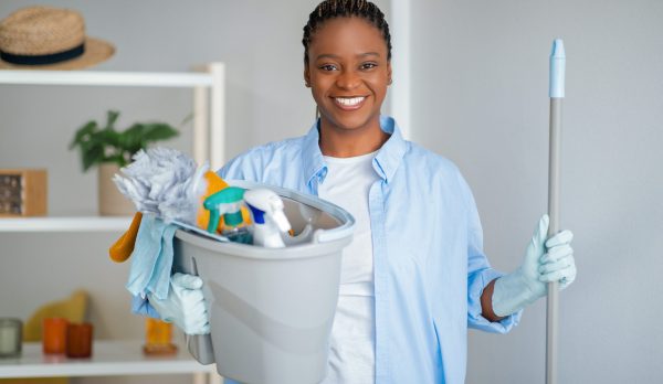 Friendly happy young african american lady in workwear maid ready for cleaning house, black woman holding basket with cleaning tools and mop, smiling at camera. Cleaning service, house-keeping